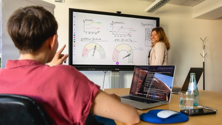 Blick über die Schulter einer kurzhaarigen Frau im Meetingraum, vorne an einem großen Screen steht eine junge Frau im Blazer, die in verschiedenen Farben Strategien und Prozesse angezeichnet hat. Die beiden unterhalten sich.