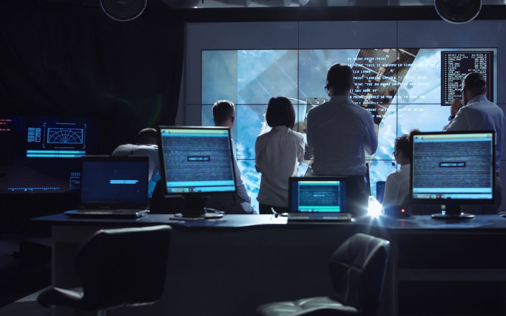 Zu sehen ist das Missionskontrollzentrum der NASA, auf einem großen Bildschirm verfolgen mehrere Menschen zwischen Computerbildschirmen eine Weltraummission.