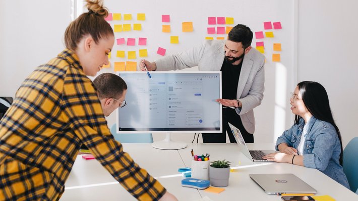 Kollegen sind um einen Konferenztisch versammelt und blicken auf einen PC-Monitor, im Hintergrund ist ein Whiteboard mit Post-Its darauf