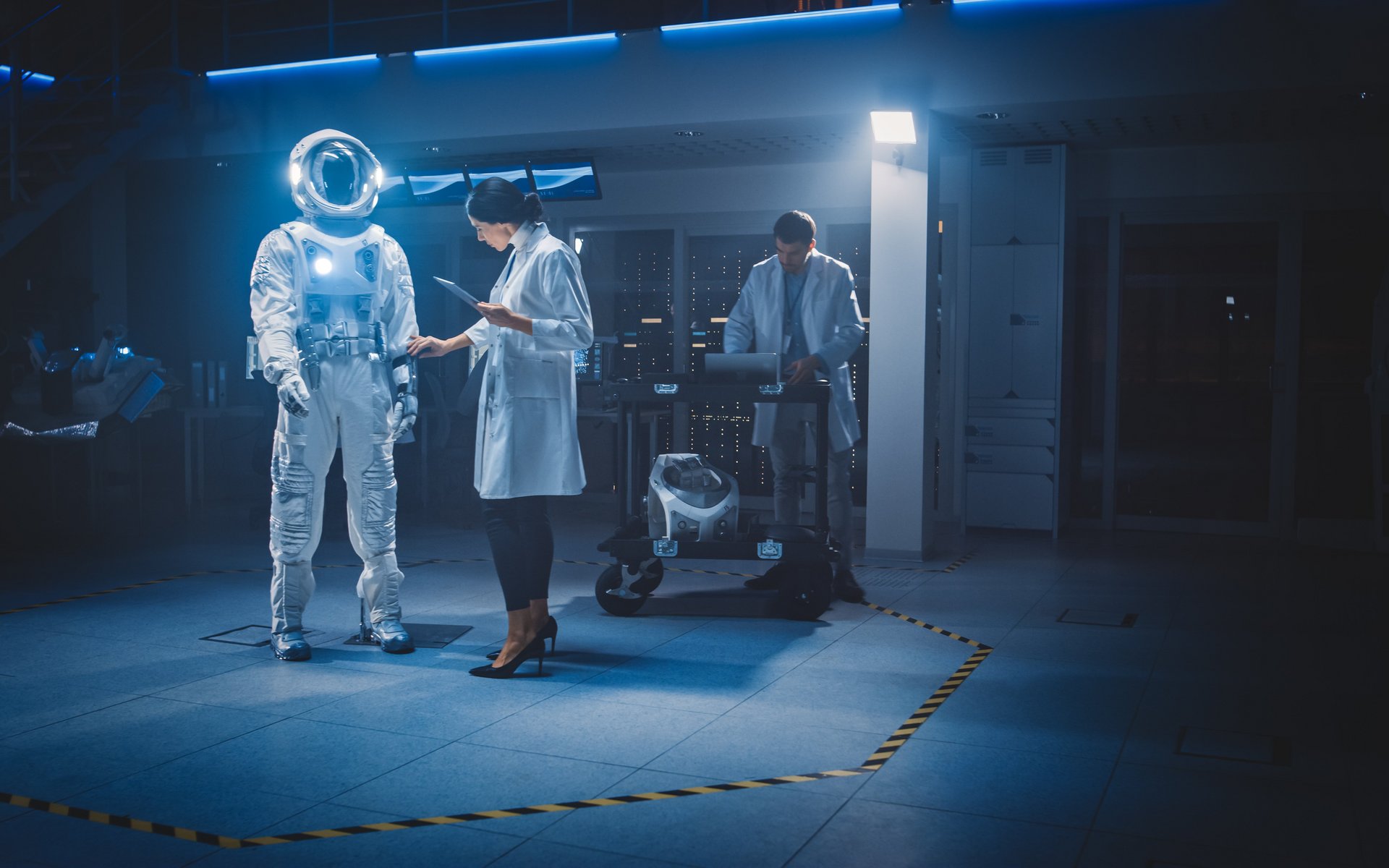In einem futuristischen Raum steht der Anzug eines Astronauten, eine Frau im weißen Kittel arbeitet daran. Ein Mann steht im Hintergrund, die Wissenschaftler konfigurieren den Raumanzug.