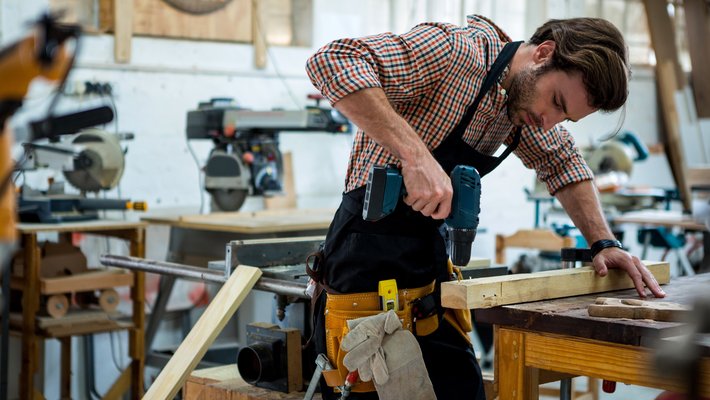 Schreiner arbeitet in seiner Werkstatt, bohrt mit Akkuschrauber in eine Holzplanke